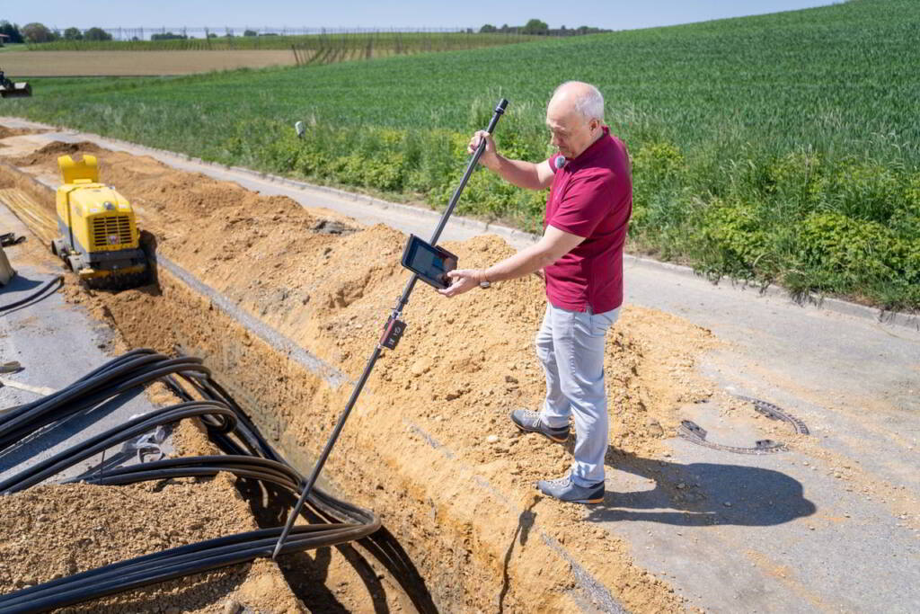 Mężczyzna z sondą pomiarową i tabletem nad rowem z czarnymi kablami, obok żółta zagęszczarka